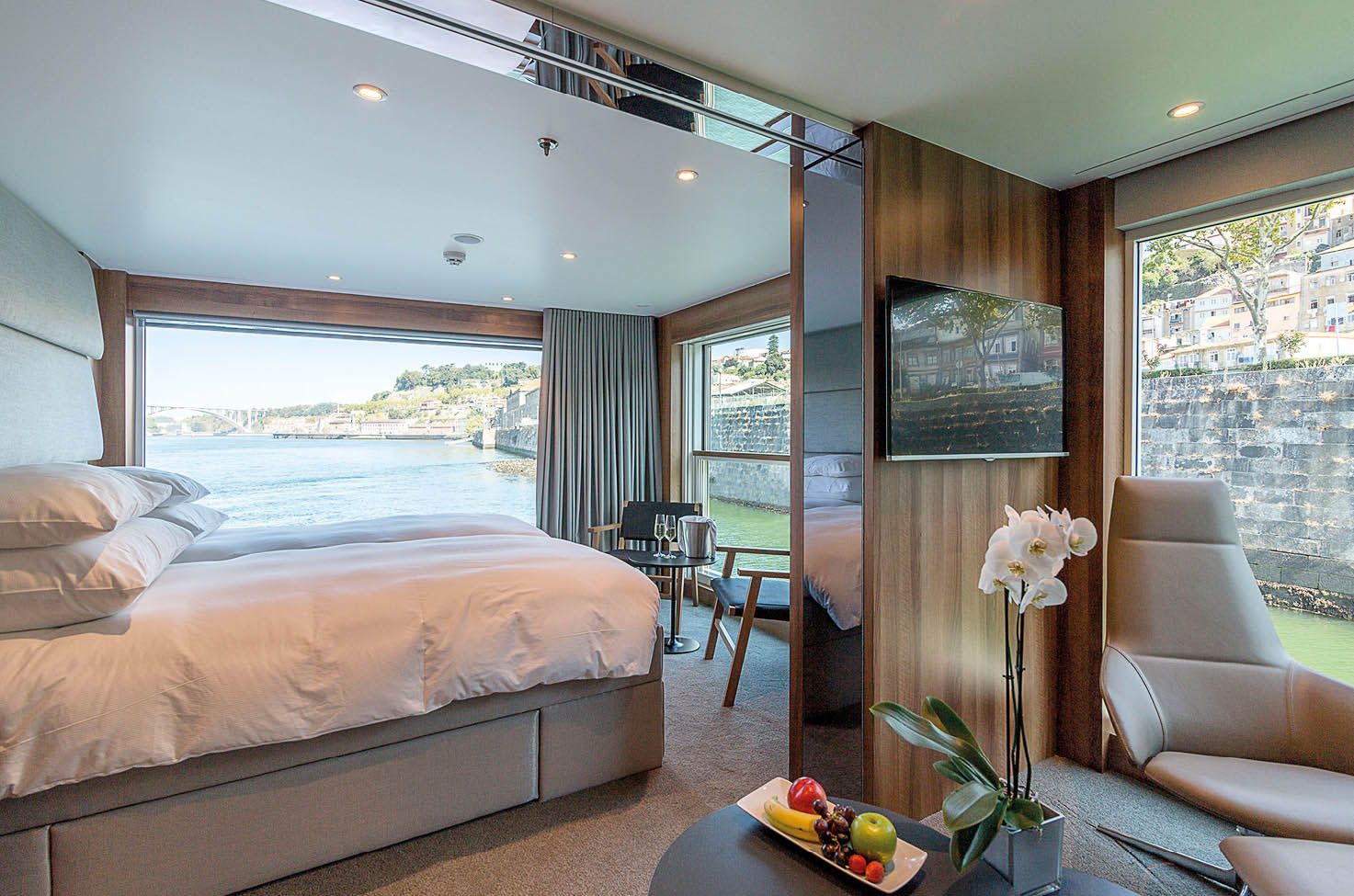 The Riverview Suite is found exclusively on the Emerald Radiance Star-Ship. A spacious suite is shown with a plush double bed, comfortable chair and large balcony-style window.