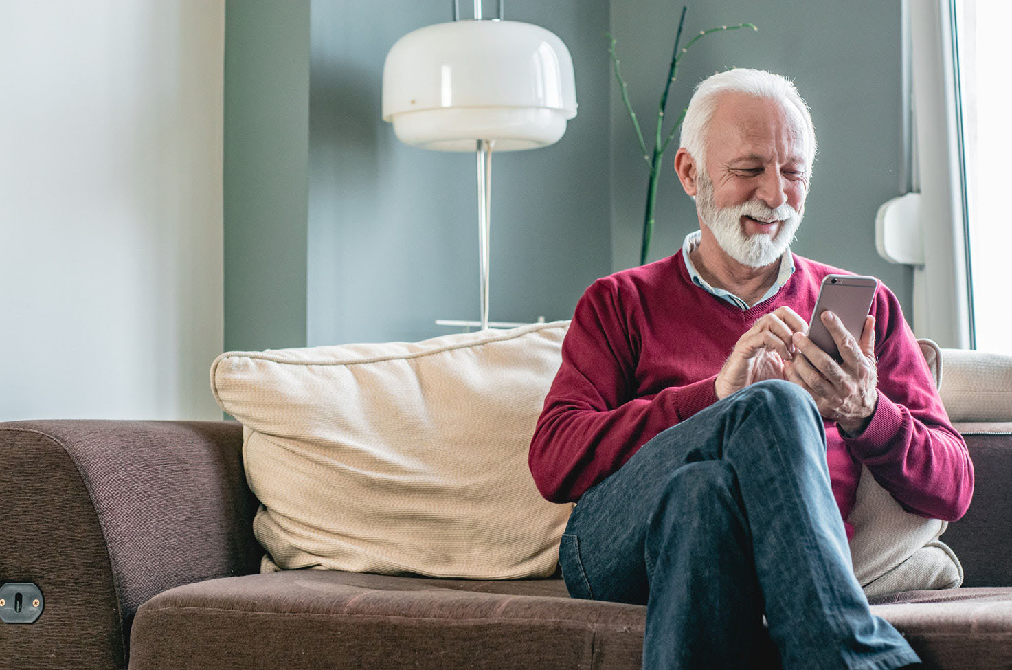 An older gentleman wearing a red jumper, sitting on a sofa, looking at a mobile phone and smiling