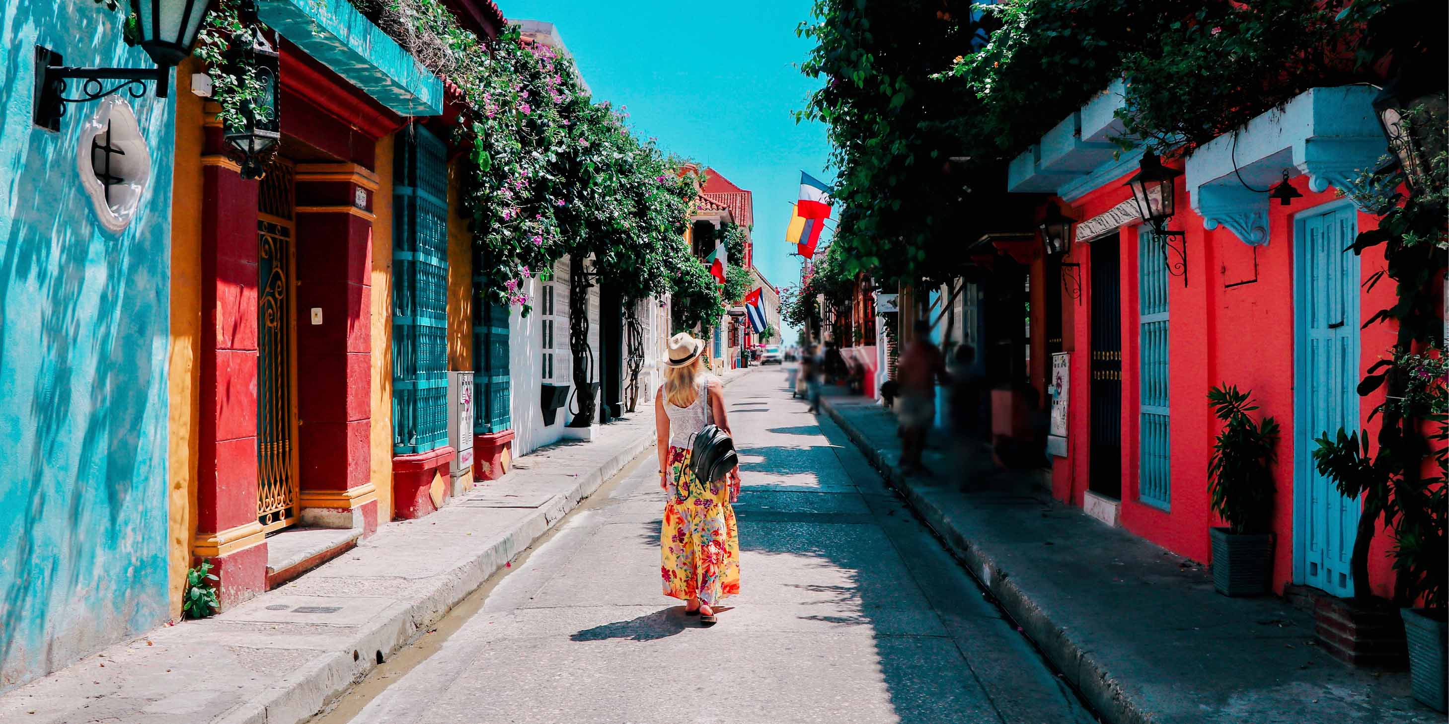 Woman walking up a street of brightly coloured buildings in the sunshine, with blue sky above, and green plants hanging from buildings