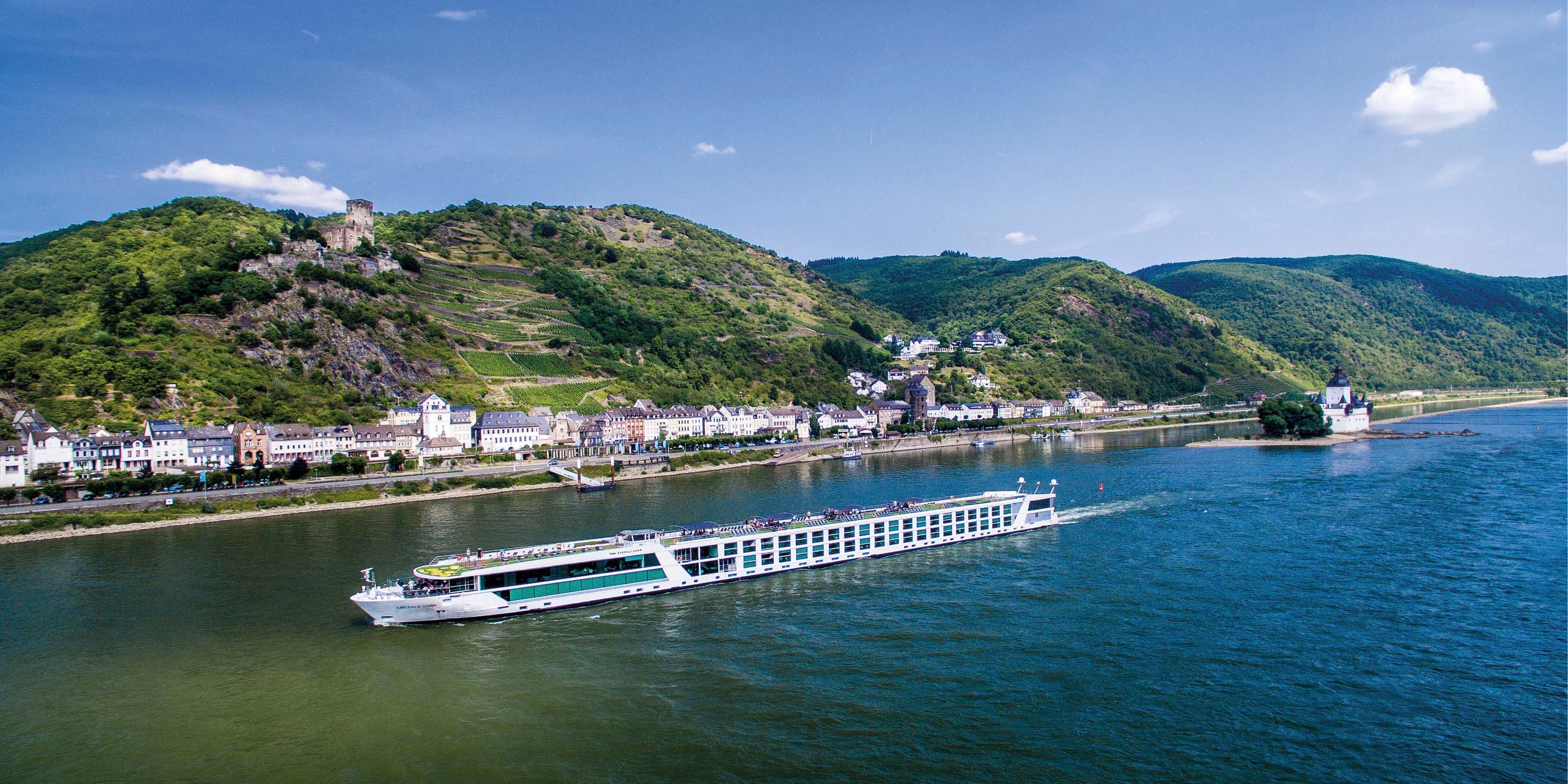 An Emerald Cruises river ship sails past a European riverbank lined with houses, with green hills in the background 