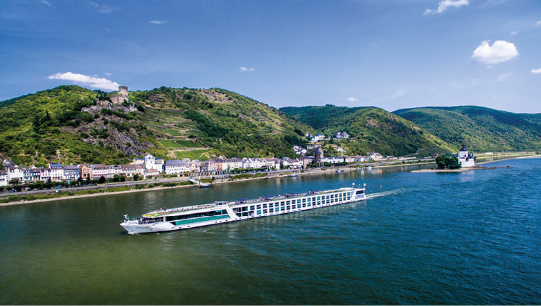 An Emerald Cruises river ship sails past a European riverbank lined with houses, with green hills in the background 