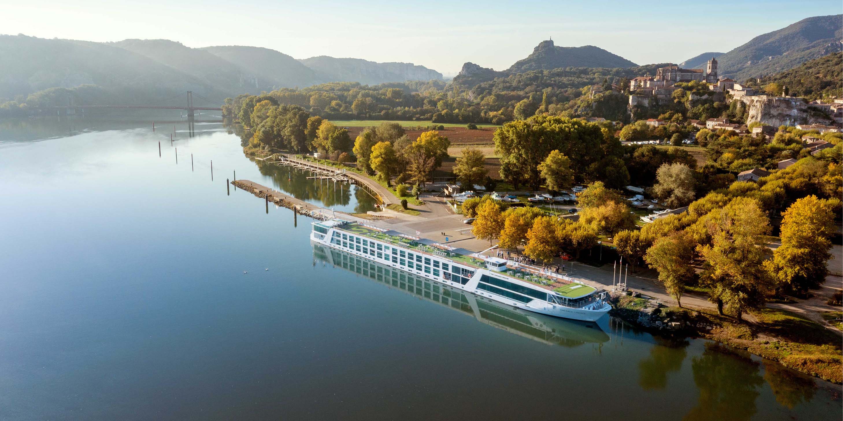 An Emerald Cruises Star-Ship is docked in Viviers, France, Green hills and a castle are seen in the background.