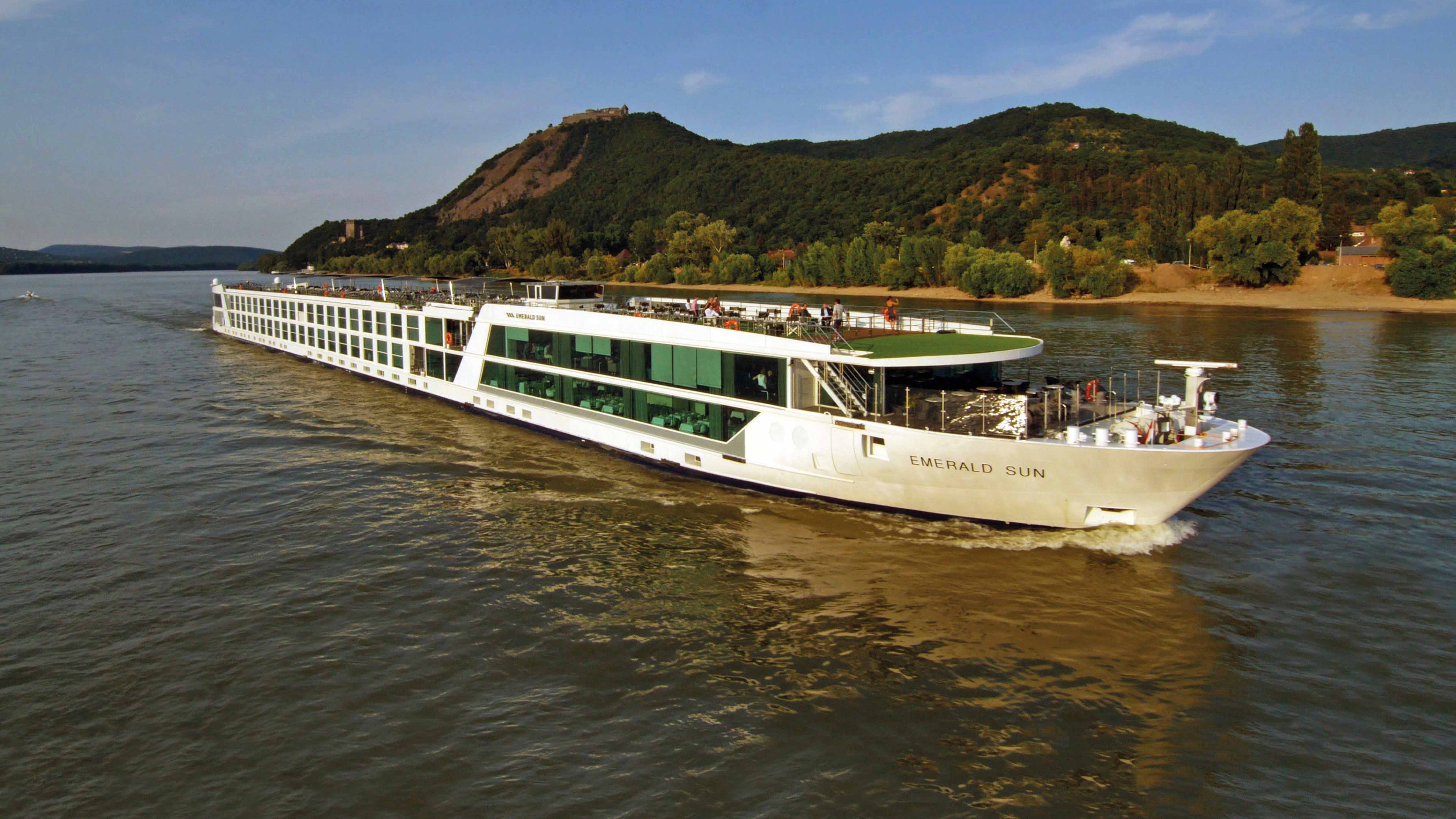 Luxury river cruise ship sailing on the rippling water past green, hilly land in Europe under a bright blue sky