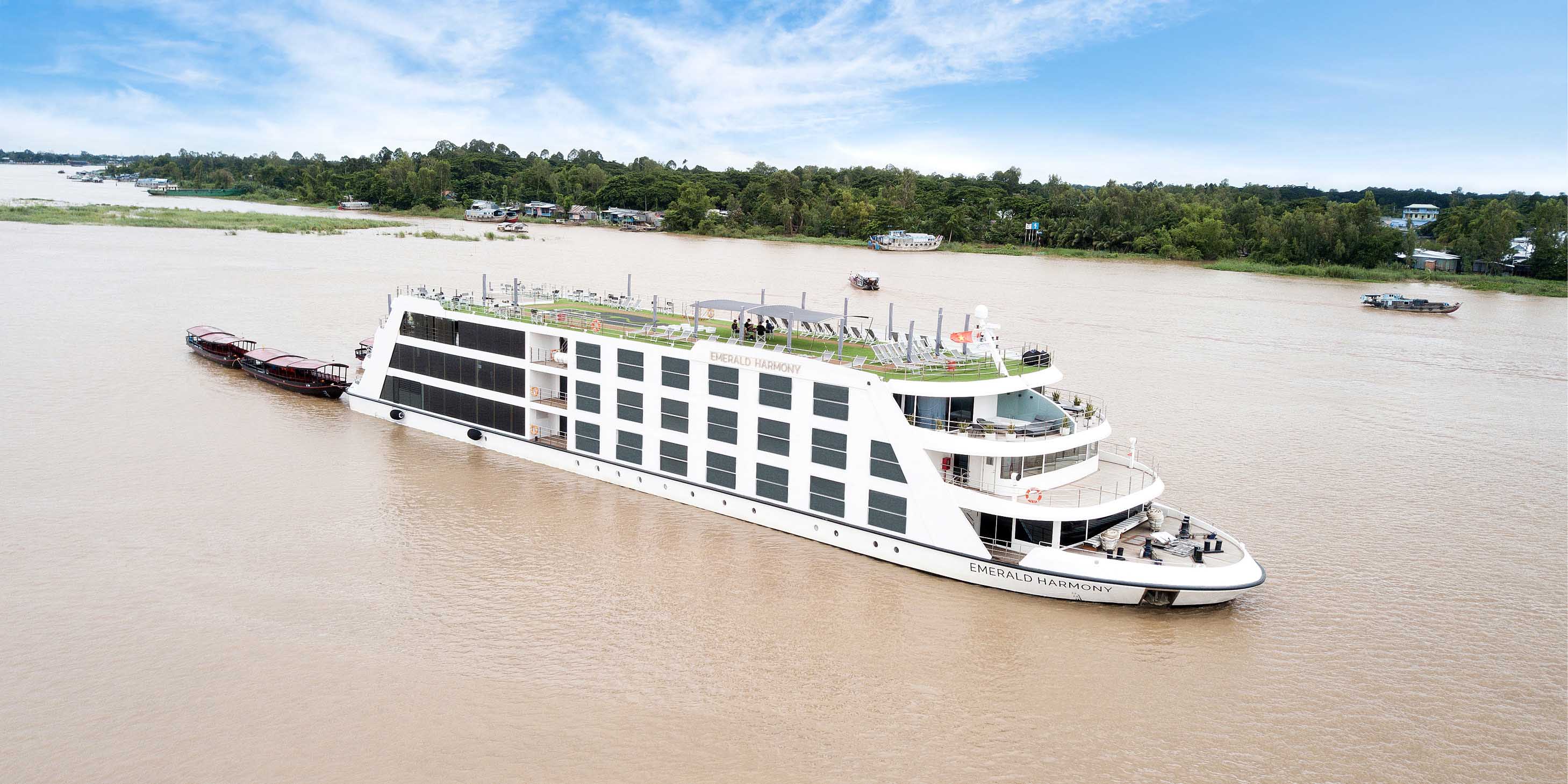 Emerald Harmony cruising along the Mekong River, with the trees along the banks and a blue cloudy sky above
