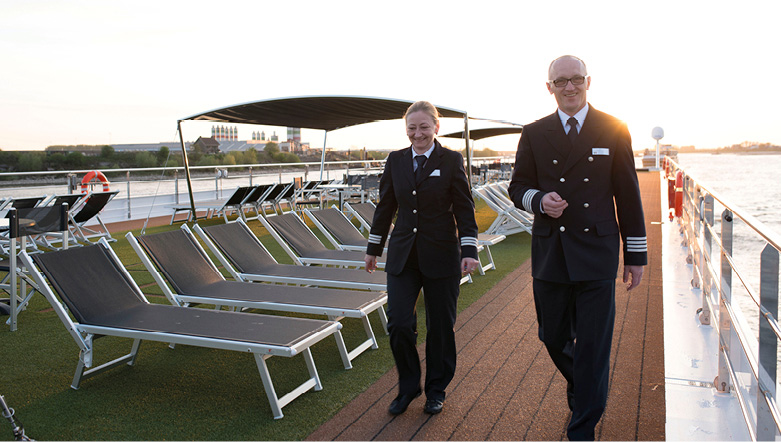 Two senior cruise ship staff walking the top deck past a row of sun loungers as the sun begins to set