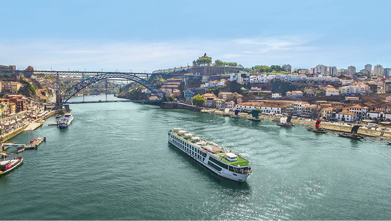 River cruise ship sailing in the middle of a river with the city of Porto on either side and an arched bridge in the background.