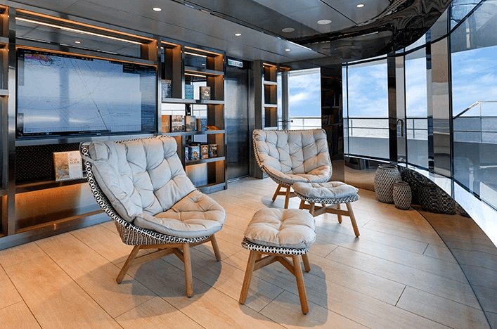 Two plush chairs and a footstool sit in a lounge with large windows in front of them. A TV and bookshelves sit behind them.