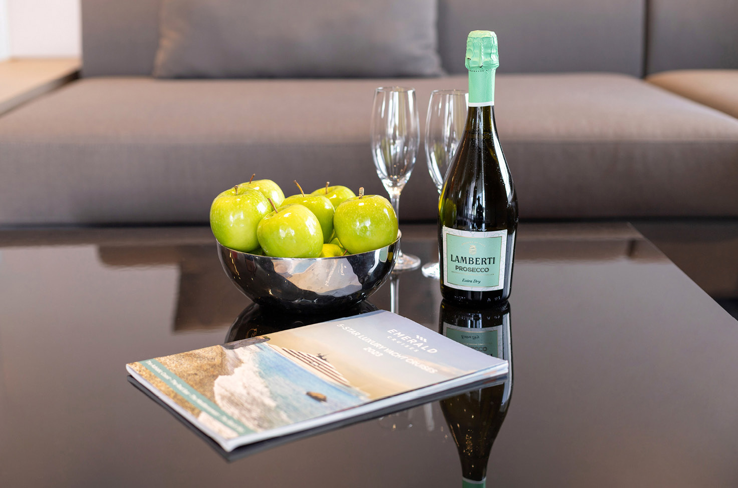 Glossy table with a bowl of green apples, a bottle of prosecco and glasses with an Emerald Cruise yacht cruises brochure 