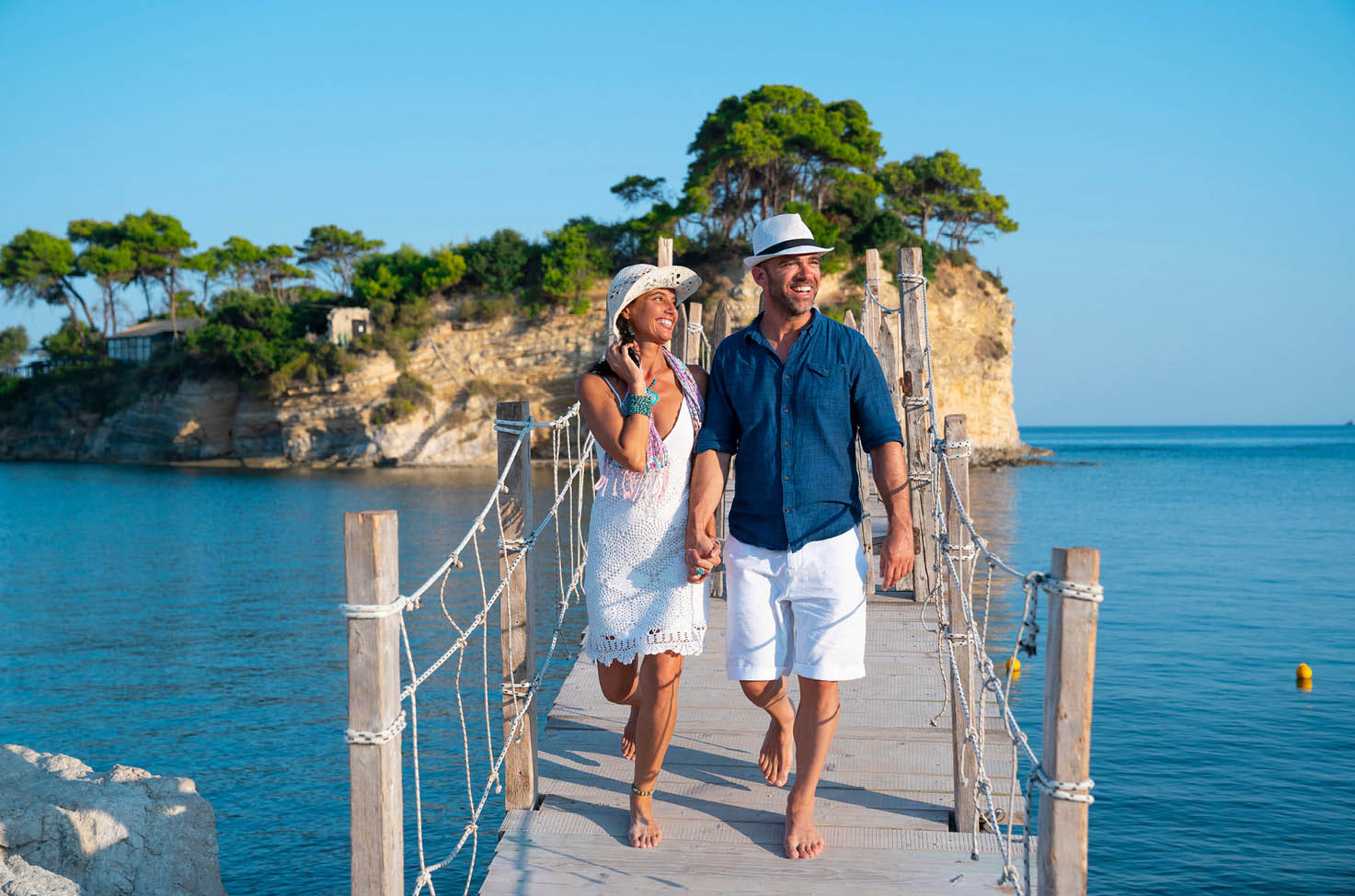  Couple holding hands wearing smart casual clothes walking across a wooden bridge across the calm blue sea with rocky island covered in greenery 