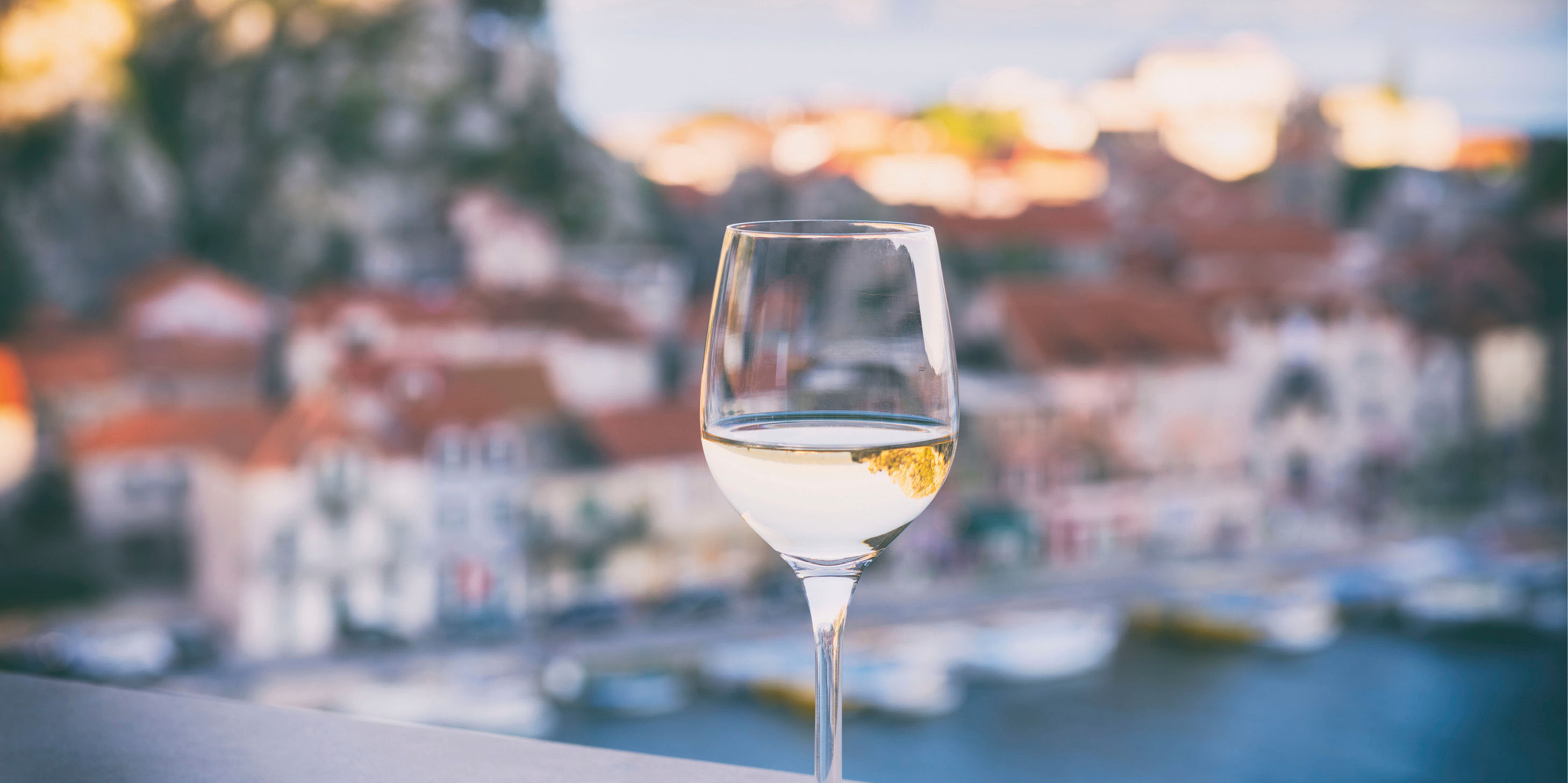 A glass of white wine in the foreground of a Mediterranean port town, which is in the shade