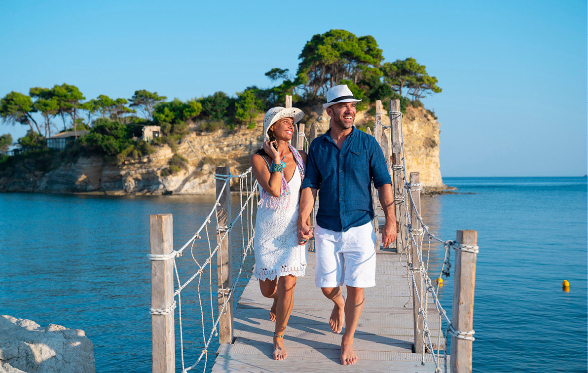 A smiling couple walk across a boardwalk on a Mediterranean island